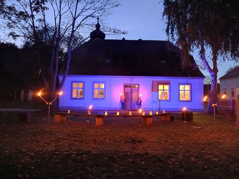 Das Schulhaus Reckahn in der Dämmerung, die Fassade ist mit lilafarbenem Licht beleuchtet.