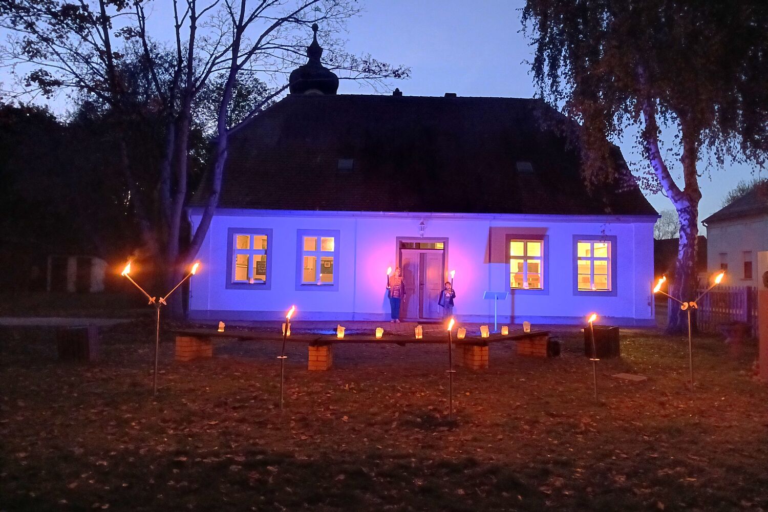 Das Schulhaus Reckahn in der Dämmerung, die Fassade ist mit lilafarbenem Licht beleuchtet.