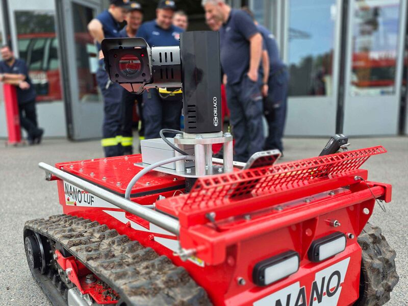 Ein ferngesteuerter Löschroboter mit aufgesetztem Kameramodul ist im Vordergrund zu sehen, während mehrere Einsatzkräfte der Feuerwehr im Hintergrund auf eine Fernsteuerung blicken.