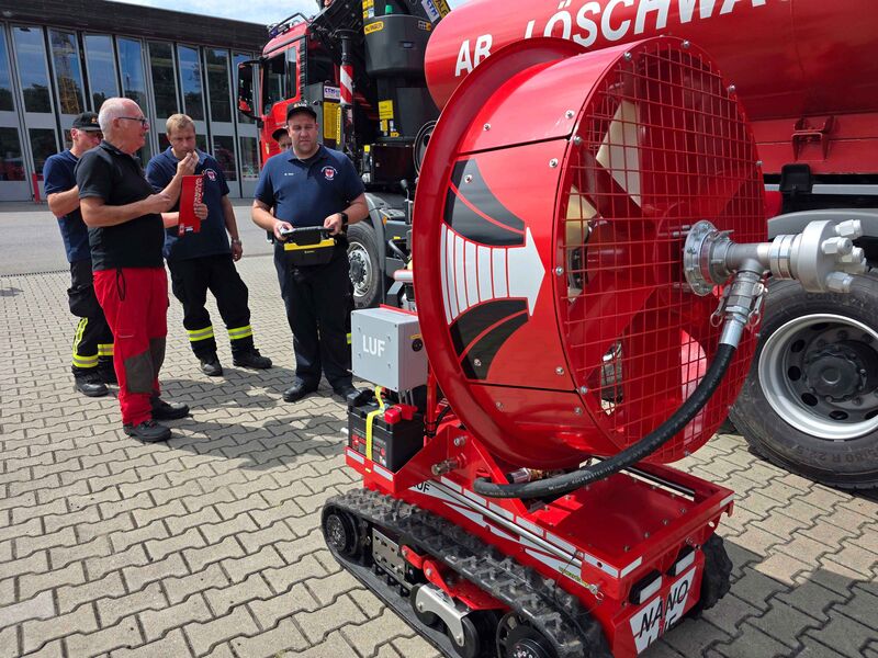 Vier Einsatzkräfte der Feuerwehr stehen hinter einem ferngesteuerten Löschroboter, während eine Person das Fahrzeug mit einer Fernsteuerung bedient.
