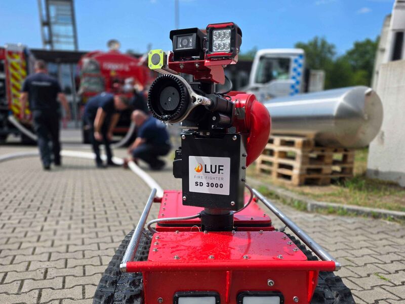 Ein ferngesteuerter Löschroboter mit angeschlossenem Schlauch ist von vorne zu sehen, während im unscharfen Hintergrund mehrere Einsatzkräfte der Feuerwehr an einem Wasserschlauch arbeiten.