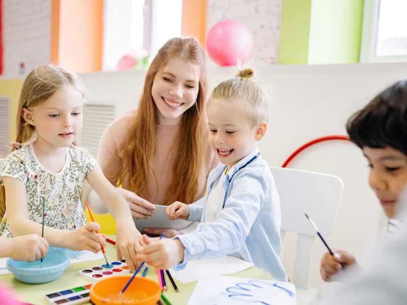 Kinder malen gemeinsam mit Wasserfarben an einem Tisch. Eine erwachsene Frau sitzt zwischen ihnen.