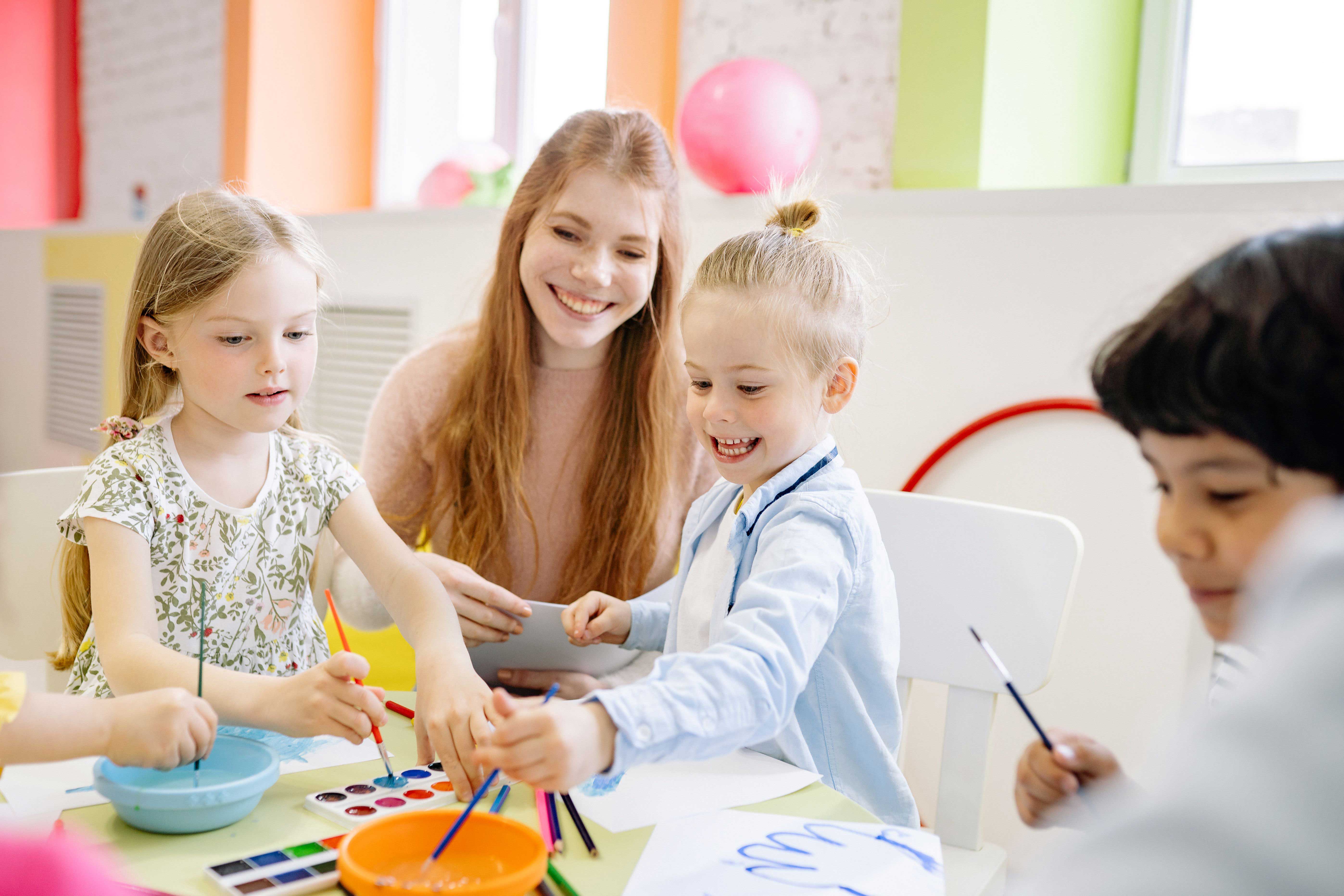 Kinder malen gemeinsam mit Wasserfarben an einem Tisch. Eine erwachsene Frau sitzt zwischen ihnen.